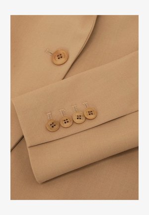 Détail d'un blazer en tissu beige avec un revers cranté et quatre boutons marron clair sur le poignet, présentant des coutures nettes et une texture lisse.