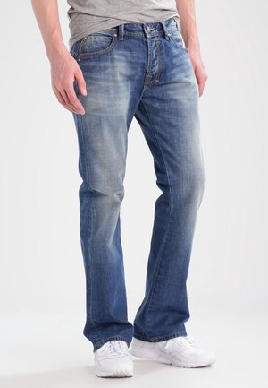 Mand iført blå slidte jeans, grå t-shirt og hvide sneakers, stående mod en ensfarvet lys baggrund.