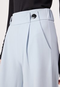 Un pantalon bleu clair présente un design ajusté avec une taille haute, des plis et deux boutons noirs à la taille pour une touche de détail supplémentaire.