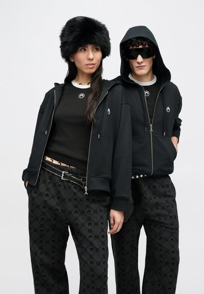 Sweats de capuz pretos com fecho de correr e bolsos, combinados com tops pretos justos e calças largas com padrão. Os acessórios incluem um chapéu de pele e óculos de sol.