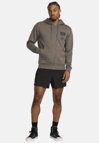 Grijze zip-up hoodie met een capuchon, voorzakken en zwart logo-patch, gecombineerd met zwarte shorts en hoge zwarte sneakers.