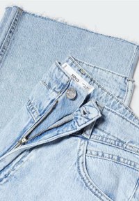 Jean en denim bleu clair avec fermeture éclair, bouton métallique et ourlet effiloché. Le tissu présente un effet délavé avec des variations de texture subtiles.