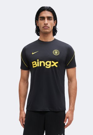 Mężczyzna stoi przodem, ubrany w czarną koszulkę piłkarską Chelsea z żółtym logo Nike i nazwą sponsora BingX.