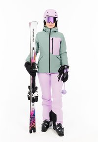 Sjezdové vybavení zahrnující zelenou a fialovou bundu se zipovou kapsou, fialové lyžařské kalhoty, černé boty a lyže s barevnými vzory.