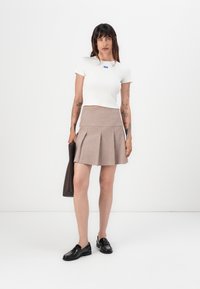 White cropped t-shirt with a blue logo, paired with a beige pleated skirt. Black shoes and a dark handbag complete the outfit.
