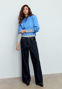 Hellblauer Strickpullover über einem schwarz-weiß gestreiften Langarmshirt, kombiniert mit weit geschnittenen dunklen Jeans und schwarzen spitzen Schuhen.