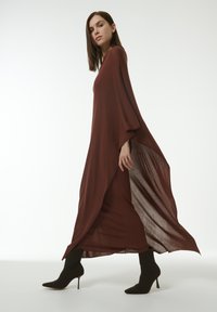 Robe longue bordeaux avec des manches larges et une fente sur le côté, fabriquée en tissu léger, associée à des bottines à talons aiguilles noires.