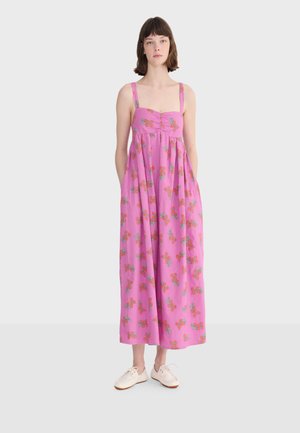 Žena oblečená v ružových, bezrukávových šatách po členky s oranžovým kvetinovým vzorom, stojaca s rukami v vreckách a béžovými topánkami na jednofarebnom pozadí.