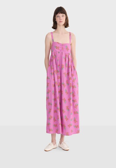 Femme portant une robe rose sans manches, longueur cheville, imprimée de fleurs orange, debout les mains dans les poches et des chaussures beiges sur un fond uni.
