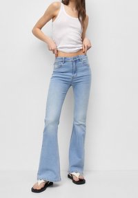 Jean évasé bleu clair en denim, avec une taille haute et un léger effet délavé, porté avec un débardeur blanc et des tongs noires.