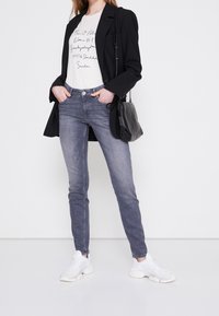 Mujer vestida con un blazer negro, una camiseta gráfica blanca, jeans skinny grises, zapatillas blancas y llevando un bolso de hombro negro.