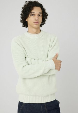 Pull en tricot côtes vert clair avec un col rond et des manches longues. Possède une coupe ajustée aux poignets et à l'ourlet. Porté sur un jean bleu foncé.