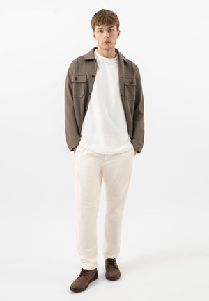 Junger Mann steht mit den Händen in den Taschen, trägt eine braune Jacke, ein weißes T-Shirt, cremefarbene Hosen und braune Schuhe vor weißem Hintergrund.