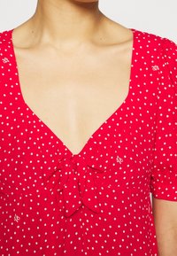 Czerwona sukienka w białe kropki, dekolt w szpic, krótkie bufiaste rękawy oraz detal w postaci wstążki na piersi. Lekki materiał i zabawny design.
