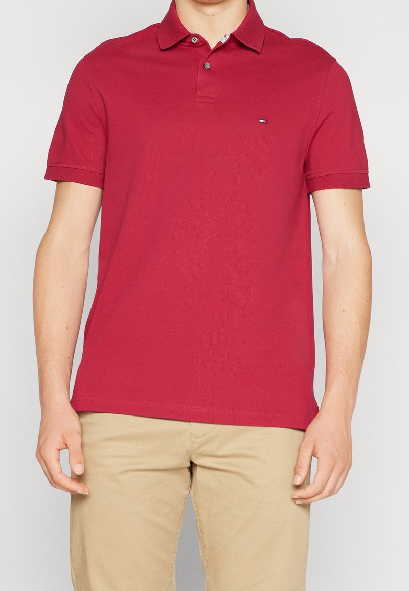 Polo en coton rouge avec col, deux boutons, manches courtes, et petit logo sur le côté gauche de la poitrine. Texture douce et coupe classique.