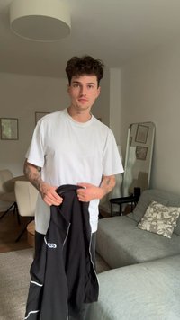 Čierna zipsová bunda s bielymi akcentmi, držiaca sa v ruke, nosená cez jednoduché biele tričko. Šedé nohavice, neformálne vnútorné prostredie s svetlými kusmi nábytku.