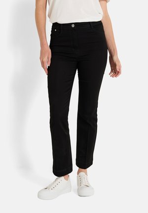 Femme portant un pantalon slim noir à chevilles avec fermeture à bouton et à glissière, associé à des baskets blanches décontractées et à un haut blanc.