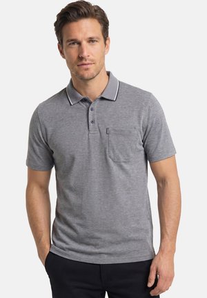 Man die een grijs polohemd met korte mouwen en een borstzak draagt, staand met één hand in zijn zak tegen een effen achtergrond.