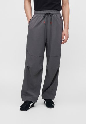 Personne portant un pantalon gris ample avec une taille élastique et un cordon, des baskets noires et une chemise noire, debout les mains dans les poches.