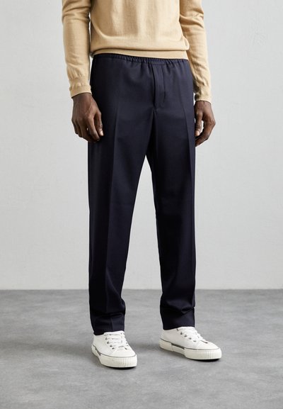 Pantaloni blu con una cintura elastica, caratterizzati da un taglio affusolato e una texture liscia, abbinati a sneakers bianche per contrasto.