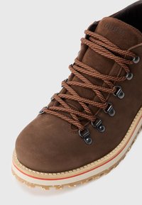 Botă de drumeție din suede maro, cu șireturi maro cu model, ochiuri metalice și un talpă bej cu o dungă roșie și un profil texturat.