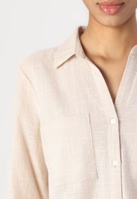Light beige shirt made of textured fabric, featuring a collar, a single chest pocket, and white buttons along the front placket.
