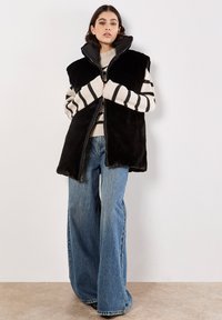 Zwarte nepbont vest met hoge kraag, ritssluiting en zijzakken, gedragen over een crèmewit en zwart gestreept sweater en blauwe wijde jeans.