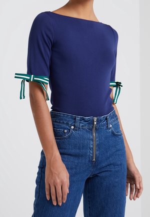 Marineblaues enges Oberteil mit weitem Ausschnitt und kurzen Ärmeln, versehen mit grünen und weißen gestreiften Krawattenakzenten an den Bündchen. Kombiniert mit hoch taillierten Jeans.