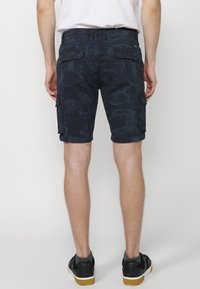 Sötétkék mintás cargo bermuda nadrágot viselő férfi, fehér pólóban, fekete sportcipőben szürke zoknival, fehér háttér felé áll.