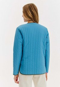 Pikowana niebieska kurtka z okrągłym dekoltem, widocznym szwem i zielonym wykończeniem na dole. Lekka, z długimi rękawami i luźnym krojem.