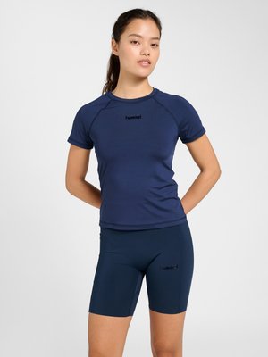 Kvinde iført navyblå atletisk t-shirt med korte ærmer og matchende knælange shorts, stående med hænderne på ryggen mod hvid baggrund.