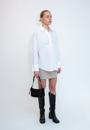 Donna con i capelli tirati indietro che indossa una camicia bianca oversized, una minigonna beige, stivali neri al ginocchio e tiene una piccola borsa nera su sfondo bianco.