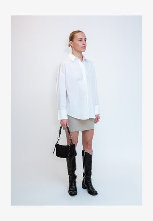 Femme aux cheveux tirés en arrière portant une chemise blanche oversize, une mini-jupe beige, des bottes noires montant jusqu’aux genoux et tenant un petit sac à main noir sur fond blanc.