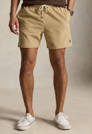 Uomo che indossa pantaloncini beige con coulisse e scarpe bianche stringate, in piedi con le mani in tasca su sfondo neutro.