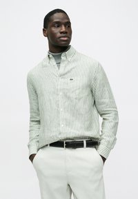 Csíkos, világoszöld és fehér anyagból készült gombos ing, mellkasi zsebbel és hímzett logóval. Világos színű nadrággal és fekete övvel viselve.