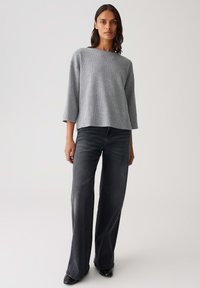 Femme portant un haut long à manches grises texturé et un pantalon en jean noir délavé à jambes larges, se tenant devant un fond clair uniforme.