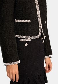 Czarna marynarka z tweedu z białą koralikową lamówką, wyposażona w usztywniane kieszenie i teksturowane rękawy. Zestawiona z pasującą czarną spódnicą.