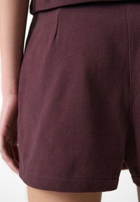 Burgundinpunaiset shortsit, joissa on tekstuuri kangas, sivutasku ja suora helma. Malli on suunniteltu istuvaksi.