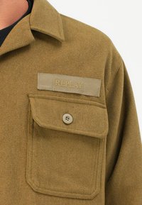 Olivenfarvet skjorte lavet af blødt stof, med en brystlomme, knaplukning og et gråt mærke med "REPLAY" broderet i beige.