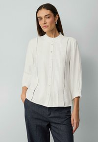 Chemise blanche à col volanté, avec des plis verticaux et des manches élastiques. Assortie à un pantalon en denim foncé pour créer un contraste.