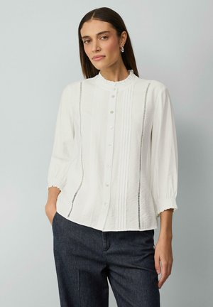 Chemise blanche à col volanté, avec des plis verticaux et des manches élastiques. Assortie à un pantalon en denim foncé pour créer un contraste.