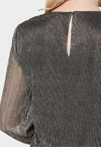 Rückansicht einer Person, die ein metallisch dunkelgraues, langärmliges Oberteil mit transparenten Ärmeln und einem kleinen vertikalen Schlüsselloch-Ausschnitt am Hals trägt.