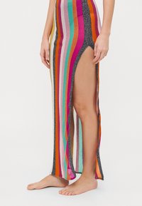 Pletené maxi šaty s jasnými, farebnými pruhmi, bočnými rozparkami a jemným lesklým textúram, v kombinácii s bosými nohami.
