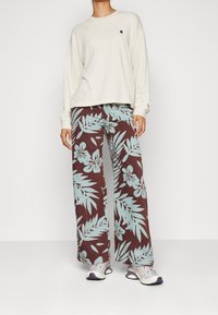 Chemise beige clair à manches longues avec un petit logo et une coupe ample, associée à un pantalon marron orné d'un motif tropical de feuilles et fleurs turquoise.