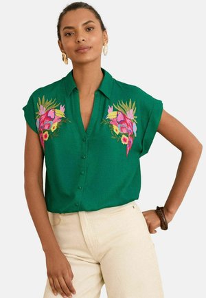 Grüne Bluse mit kurzen Ärmeln, ausgestattet mit lebhaften gestickten Tukanen und floralen Designs auf der Brust. Knopfverschluss vorne.