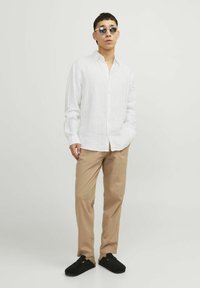 Weißes Leinenhemd mit langen Ärmeln, beiges Hosen und schwarze Slipper. Modell trägt runde Sonnenbrillen. Schlichter Hintergrund.