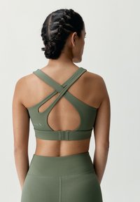 Zelená sportovní podprsenka s překříženými bandážemi a detailem výřezu. Hladký, pružný materiál v kombinaci s vysoce pasovými legínami v odpovídající barvě.