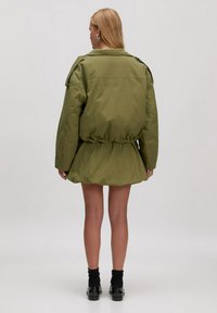 Olijfgroene parka jas met een getailleerde taille, lange mouwen en een uitlopende zoom. Heeft een grote kraag en zijzakken. Soepele textuur.