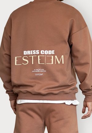 Man wearing brown sweatshirt and pants, sweatshirt back text reads "DRESS CODE ESTEEM, HIGH STANDARD STREETWEAR" in white and beige letters.
