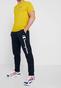 Yellow short-sleeve t-shirt with logo, navy sweatpants featuring white text, and white sneakers with red and blue accents.
